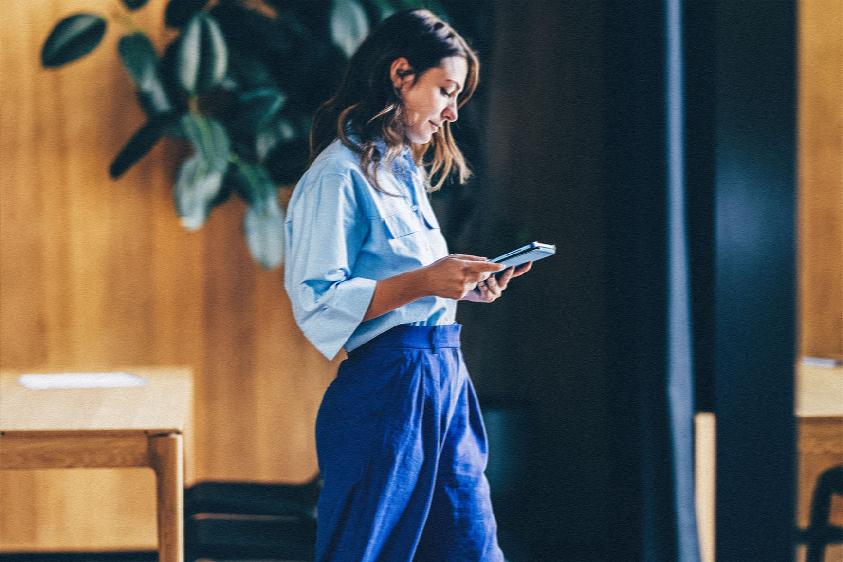 A woman in loose casual attire holds a tablet device and gazes down on it.
